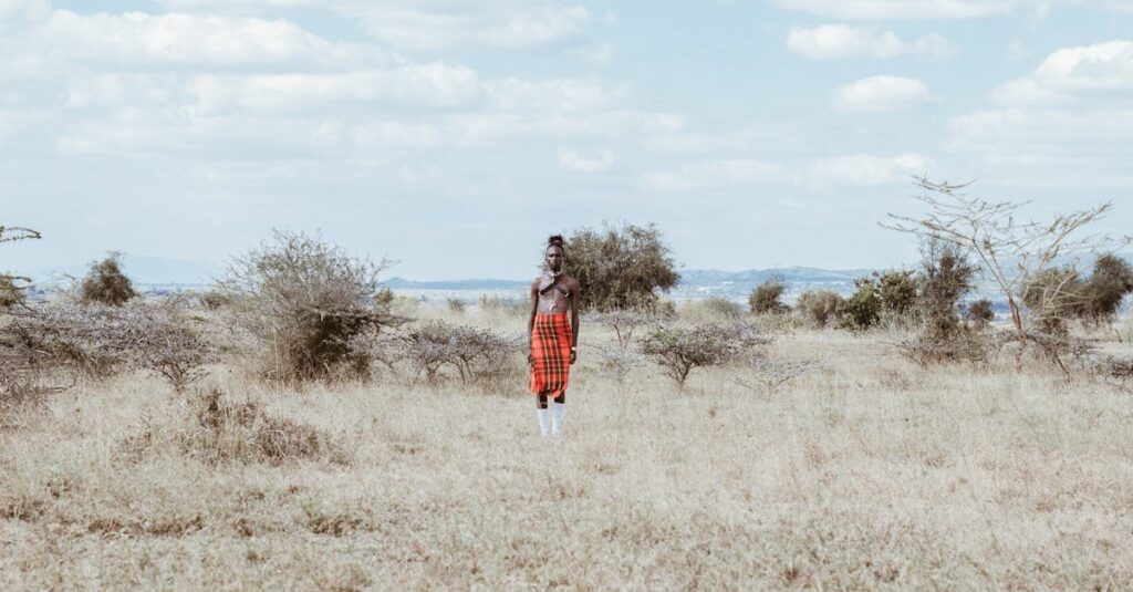 A person in traditional Kenyan attire stands in a vast grass field under a cloudy sky.